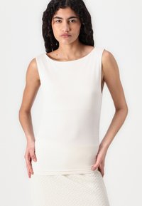 Αμάνικο κρεμ τοπ από μαλακό ύφασμα, με ευρύτερη λαιμόκοψη και εφαρμοστή σιλουέτα. Γυναίκα στέκεται με τα χέρια στη μέση.