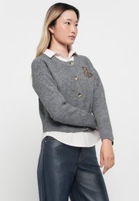 Cardigan tricoté gris avec des poignets côtelés, doté de boutons ronds et d'un détail brodé d'ours sur le côté gauche. Porté sur une chemise blanche.