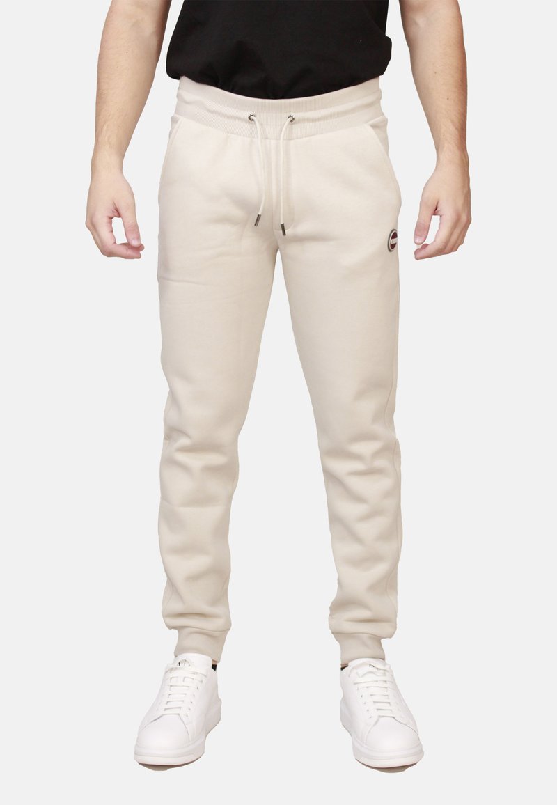 Pantaloni della tuta beige con vita regolabile, polsini a coste, tasche laterali e un piccolo logo sul coscio sinistro. Abbinati a sneakers bianche.