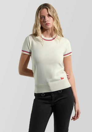 Donna bionda che indossa un maglione bianco a maniche corte con bordi rossi e blu e pantaloni neri, in piedi davanti a uno sfondo grigio chiaro.