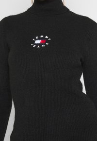 Czarny dziany sweter z golfem o prążkowanej strukturze, z okrągłym białym haftem z napisem "TOMMY JEANS" z czerwonymi i niebieskimi akcentami.