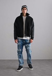 Schwarze Fleece-Kragenjacke mit Reißverschluss, kombiniert mit verwaschenen blauen Jeans und schwarzen Sneakers. Eine schwarze Mütze rundet das Outfit ab.
