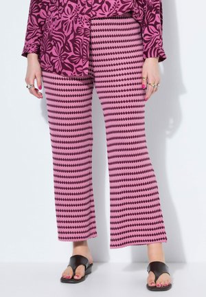 Person iført lyserøde og mørkelilla mønstrede bukser og blomsterprintet top med sorte åbne sandalier, der står ved en hvid væg.