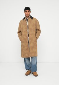 Tan, knee-length coat with a button front, large collar, and side pockets. Worn over a gray and checkered shirt with blue jeans and brown shoes.