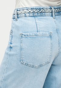 Φωτεινό γαλάζιο τζιν παντελόνι με πίσω τσέπη και πλεκτή ζώνη από τζιν, συνδυασμένο με λευκή μπλούζα.