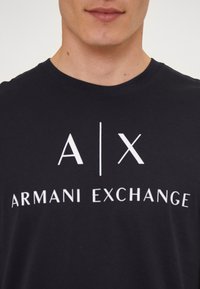 Juodas medvilnės marškinėliai su baltu grafinio logotipo užrašu "A|X" virš "ARMANI EXCHANGE" teksto, apvaliu kaklu ir trumpomis rankovėmis.