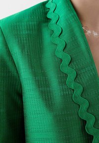 Green textured fabric blazer featuring a wavy accent trim along the neckline, with subtle geometric patterns creating depth.