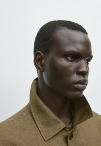 Brown linen jacket with a high collar, featuring a button closure and textured fabric. Model has short, closely cropped hair.