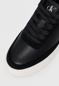 Sneaker nera in pelle e suede, con superficie texturizzata, punta rotonda, suola bianca e lacci neri con etichetta brandizzata sulla linguetta.