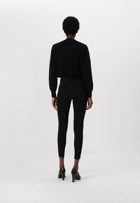 Suéter negro de punto con hombros caídos, combinado con leggings negros. Lleva zapatos de tacón alto negros. Silueta ajustada y suave.