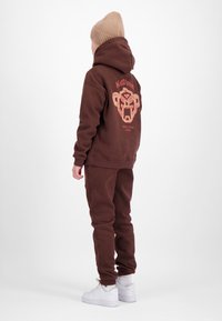 Persoon met een bruine hoodie en bijpassende joggers met grafisch ontwerp op de achterkant, beige gebreide muts, witte sneakers, staand tegen een witte achtergrond.