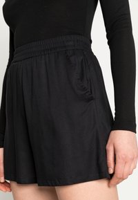 Schwarze, leichte Shorts mit elastischem Bund, seitlichen Taschen und einem lockeren Schnitt. Glatte Textur und gesäumte Kanten.