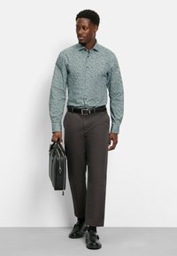 Lichtblauw patroon shirt, lange mouwen, knoopsluiting. Gepaard met een bruine broek en zwarte schoenen. Houdt een zwarte leren aktetas vast.