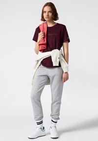 Bordeaux katoenen t-shirt met geborduurd logo, grijze sweatpants, witte sneakers met groene accenten, en een crèmekleurige sweatshirt om de taille geknoopt.