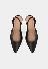 Musta teräväkärkinen slingback-läskipohjainen, jossa on säädettävät soljenuurit ja pehmeät vaaleanruskeat pohjat, aseteltuna vierekkäin valkoista taustaa vasten.