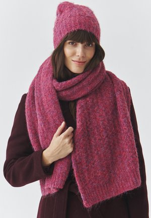 Roosa kootud beanie ja sallikomplekt, millel on tekstureeritud muster. Materjalid näivad olevat pehmed ja soojad, täiendades tumedat mantlit.