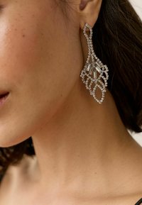 Pendientes grandes en tono plateado con un intrincado diseño de hojas, adornados con piedras de strass transparentes y acentos rectangulares, colgando elegantemente de la lóbulo.