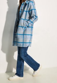 Blauer und grauer karierter Wollmantel mit Kapuze, Knopfverschluss und Seitentaschen, kombiniert mit blauer Jeans und weißen Turnschuhen.