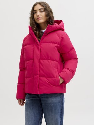 Kvinde med langt bølget hår iført en lys pink polstret jakke med hætte og blå jeans, stående med den ene hånd i lommen.