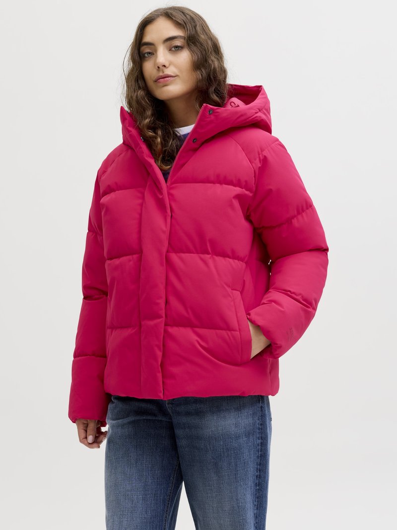 Woman with long wavy hair wearing a bright pink padded jacket with hood and blue jeans, standing with one hand in pocket.