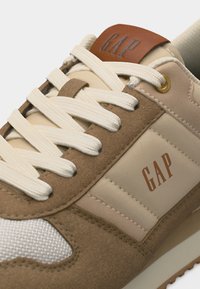 Μπεζ και καφέ αθλητικό παπούτσι με λευκά κορδόνια, πλέγμα στη μύτη και λογότυπο «GAP» στην πλευρά και τη γλώσσα, με λεπτομέρεια από χρυσό μεταλλικό οφθαλμό.