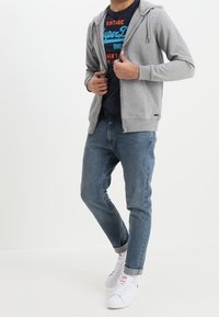 Grå zip-up hoodie med snørelukning over en marineblå t-shirt med blå og rød grafisk tekst, parret med lyseblå jeans og hvide sneakers.