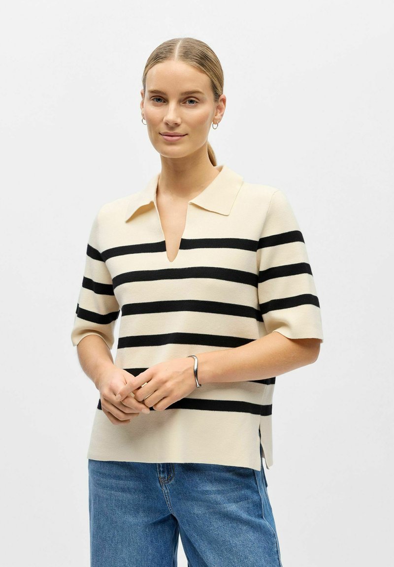 Femme à la peau claire et aux cheveux blonds attachés, portant un haut beige à manches courtes avec des rayures horizontales noires et un jean bleu.