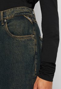 Close-up van de donkerblauwe denimjeanszak met zichtbare stiksels en een klein Replay-merklabel, gedragen met een zwart longsleeve shirt.