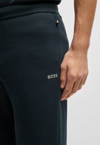 Mørkeblå teksturerte bukser med et brodert "BOSS"-logo på låret og en liten metallisk detalj nær midjen.