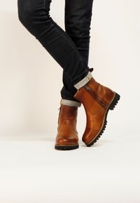 Botines de cuero marrón con acabado texturizado, cremallera lateral y suela de goma negra, combinados con jeans oscuros con puños.