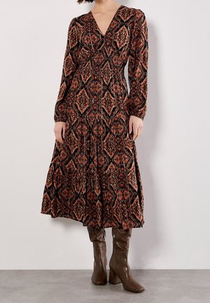 Donna che indossa un vestito a maniche lunghe con scollo a V con un motivo geometrico marrone e nero e stivali marroni con tacco fino al ginocchio.