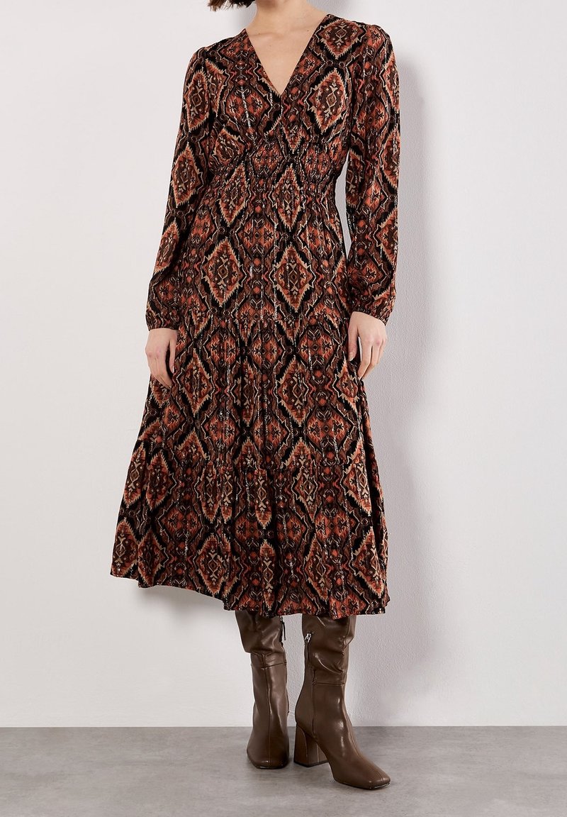 Femme portant une robe à manches longues avec un col en V, un motif géométrique marron et noir, et des bottes marron à talons montant jusqu'au genou.