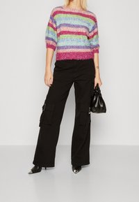 Meerkleurige, pluizige trui met horizontale strepen in roze, groen, blauw en paars; gecombineerd met zwarte cargobroek en zwarte pumps met puntige neus.