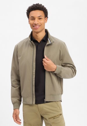 Jeune homme souriant portant une veste légère marron clair à fermeture éclair, un polo noir et un pantalon cargo beige avec une poche zippée, sur fond blanc.