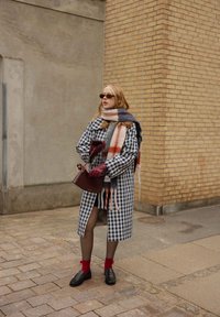 Manteau ceinturé à carreaux bleu et blanc ; écharpe rayée grise, orange et marine ; sac à main bordeaux ; chaussettes rouges ; mocassins noirs sur pavés.
