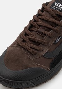 Brązowe zamszowe sneakersy z czarnymi gumowymi akcentami, teksturowanym materiałem i płaskimi sznurowadłami. Posiadają logo Vans na języku oraz wyściełany kołnierz.