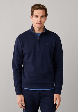 Young man wearing a navy blue quarter-zip sweatshirt over a light blue shirt, standing against a plain gray background.