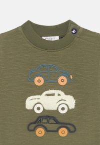 T-shirt verde oliva con tre auto giocattolo ricamate in blu, bianco e blu scuro, con ruote arancioni e dettagli texture sulla macchina bianca.