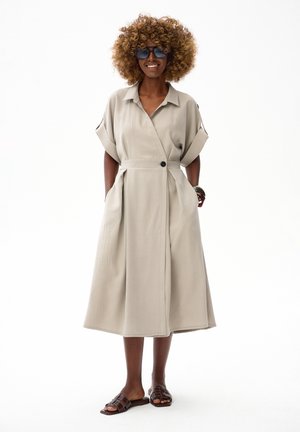 Frau mit lockigem Haar, die dunkle Sonnenbrillen trägt und ein beiges Midi-Kleid mit kurzen Ärmeln, Seitentaschen und braunen Sandalen zum Hineinschlüpfen anhat.