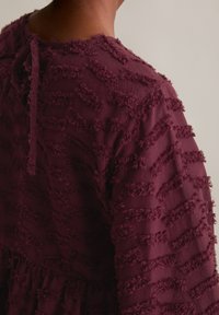 Burgunderfarbenes Kleid aus strukturiertem Stoff mit horizontalen, wellenförmigen Fransenstreifen; runder Ausschnitt mit einer Bindung im Rücken.