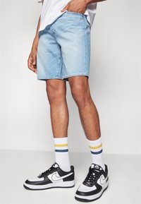 Henkilö, joka pukeutuu vaaleansiniisiin farkkushortseihin, valkoisiin sukkiin, joissa on keltaisia ja sinisiä raitoja, sekä musta-valkoisiin Nike Air Force 1 -lenkkareihin.