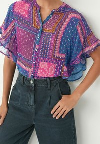 Blusa a maniche corte colorata e fantasia, caratterizzata da tonalità di viola, blu e rosa, con una chiusura frontale a bottoni e un design morbido e flessuoso. Indossata con jeans scuri.