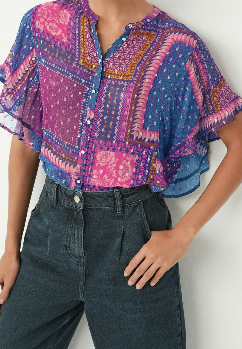 Blusa a maniche corte colorata e fantasia, caratterizzata da tonalità di viola, blu e rosa, con una chiusura frontale a bottoni e un design morbido e flessuoso. Indossata con jeans scuri.