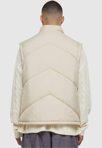 Ljusbeige pufferväst med hög krage, chevronquirat design och en mjuk textur. Kombinerad med en kabelstickad tröja under.