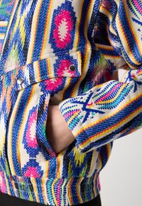 Hand in de zak van een jas met kleurrijke, ingewikkelde geometrische patronen in blauw, roze, geel en groen op een beige stof.