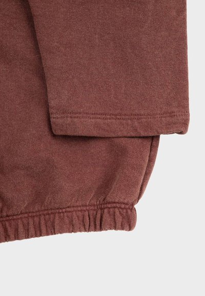 Nahaufnahme eines braunen Hemds mit langen Ärmeln und elastischem Bund aus weichem Strickstoff vor hellem Hintergrund.