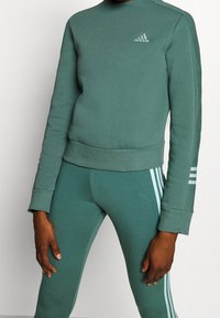 Conjunto de sudadera verde y leggings, hecho de una tela suave, que presenta un cuello alto, puños acanalados y tres rayas blancas en el lado.