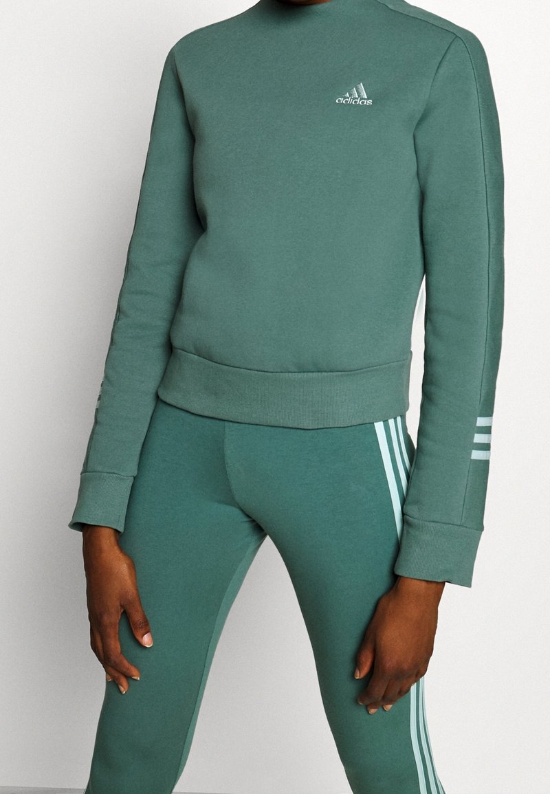 Conjunto de sudadera verde y leggings, hecho de una tela suave, que presenta un cuello alto, puños acanalados y tres rayas blancas en el lado.
