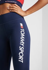Námornické modré legíny vyrobené z pružného materiálu, s bílým logem "TOMMY SPORT" na boku a přiléhavým vysokým pasem.