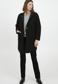 Fekete, texturált külső kabát nagy kapucnival, pattintós gombokkal és két elülső zsebbel. Egy világosszürke felső és fekete nadrág felett viselve.